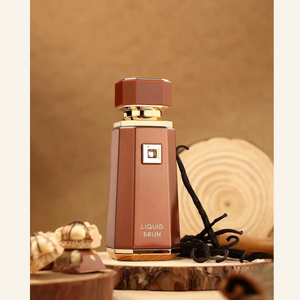 A faceted brown perfume bottle labeled 'LIQUID BRUN' with gold trim, displayed on a wood slice with vanilla beans and chocolates against a warm brown background.