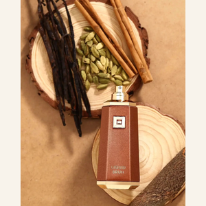 A brown rectangular perfume bottle labeled 'LIQUID BRUN' with a gold spray top, styled on wood slices with cinnamon sticks, cardamom seeds, and vanilla beans on a tan background.